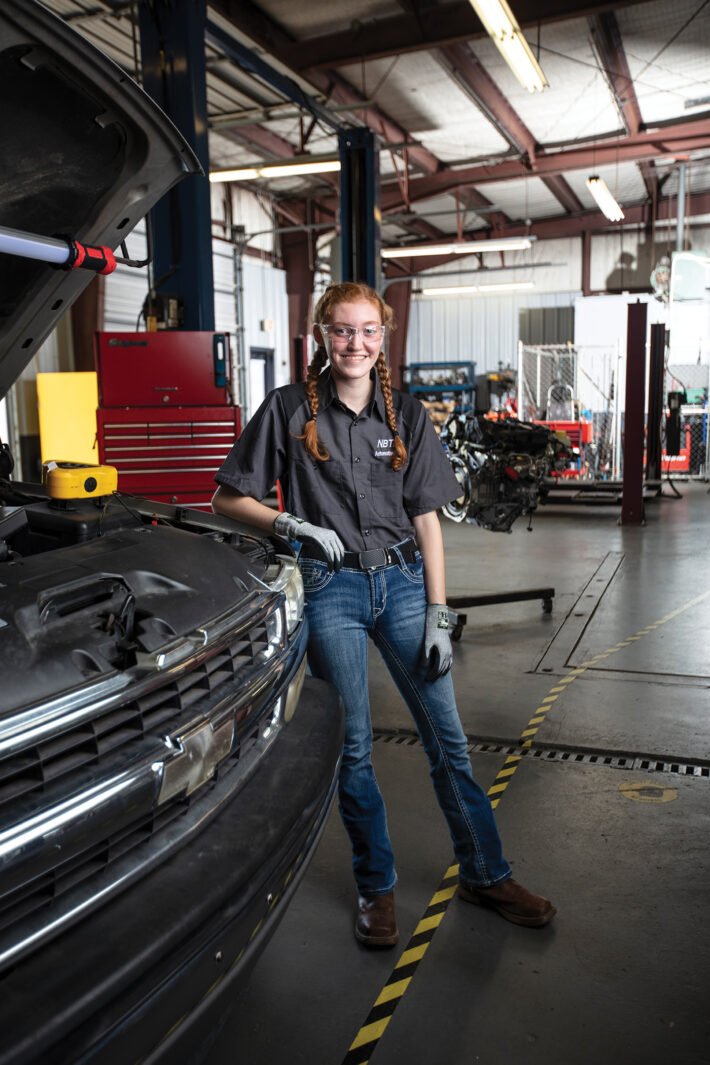 female automotive technician standing in front of open car hood with protective goggles on