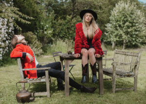 Mary Bragg and Willow Robinson of Outpost Drive in red and black outfits posing on outdoor furniture